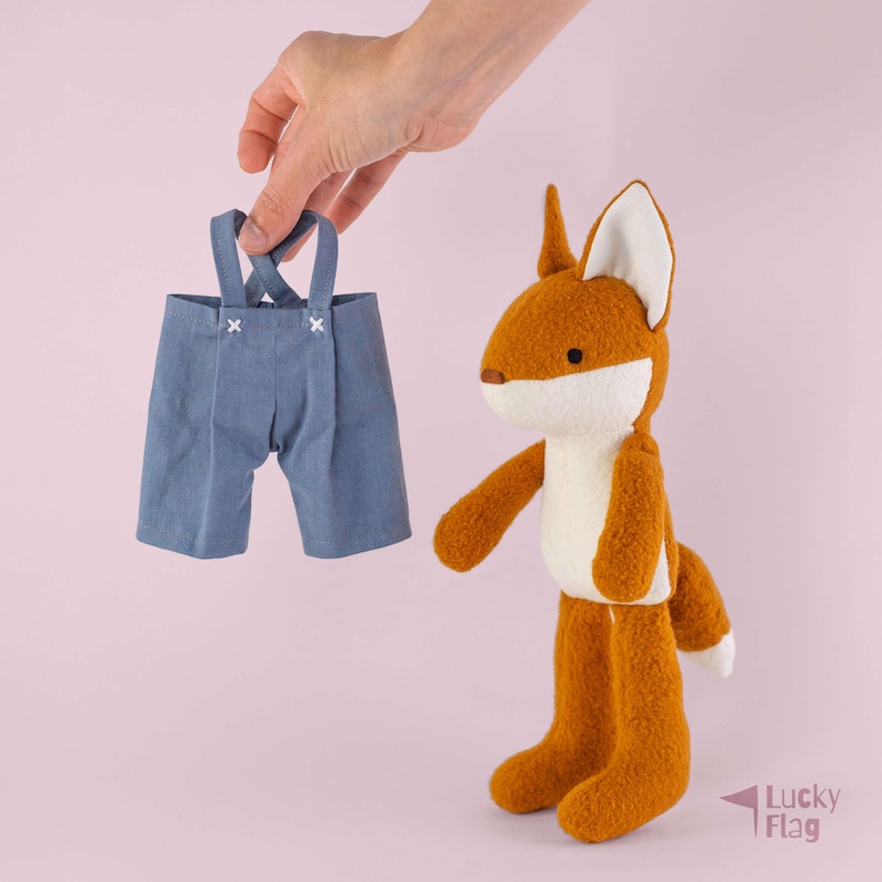 May include: A plush fox toy with orange fur, a white chest, and white-tipped ears, standing upright. Beside it are blue fabric shorts with suspenders featuring white cross-stitch detailing at the attachment points on the front waistband.