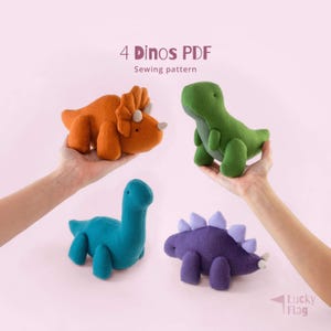 Peut inclure: Quatre peluches de dinosaures colorées : un tricératops orange, un T-Rex vert, un Brontosaure bleu et un Stégosaure violet. Le texte "4 Dinos PDF Sewing pattern" est en haut. Le logo Lucky Flag est en bas à droite.
