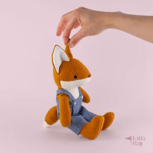 May include: A plush fox toy with orange fur, a white chest, and white-tipped ears. The fox is wearing blue overalls with white stitched details. The toy has a small brown nose and black eyes, and it is shown in a seated position with a tail.