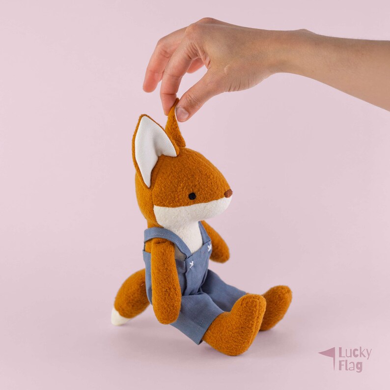 May include: A plush fox toy with orange fur, a white chest, and white-tipped ears. The fox is wearing blue overalls with white stitched details. The toy has a small brown nose and black eyes, and it is shown in a seated position with a tail.