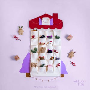 Puede incluir: Un calendario de adviento de tela color crema con forma de casa, con tejado rojo y 24 bolsillos. Los bolsillos contienen pequeños peluches y elementos decorativos. El texto "*Plushies not included" está en la parte inferior.
