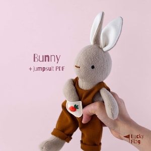 Könnte beinhalten: Ein beiges Plüschhasenspielzeug mit langen, weiß gefütterten Ohren, das eine braune Latzhose mit einer kleinen weißen Tasche und einer aufgestickten orangefarbenen Karotte trägt. Der Text Bunny + jumpsuit PDF steht links, Lucky Flag unten rechts.