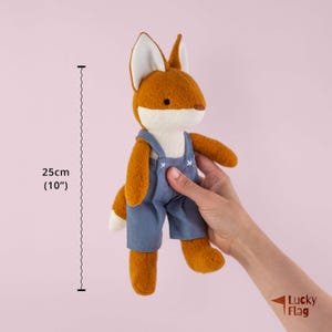 May include: A plush fox toy with orange fur, a white muzzle, and white-tipped ears, wearing blue overalls with white stitched details. A measurement graphic indicates a height of 25 cm or 10 inches for this soft, stuffed animal figure.