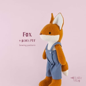 Puede incluir: Un peluche de zorro con pelaje naranja, detalles blancos y un peto azul. El zorro tiene hocico y orejas blancos. El texto en la imagen dice "Fox + pants PDF Sewing pattern". El juguete está sobre un fondo rosa.
