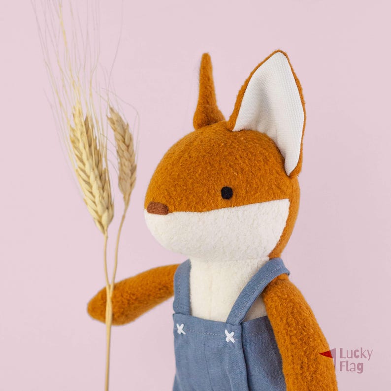May include: A plush fox toy with orange fur, a white muzzle, and white-lined ears. The fox wears blue denim-style overalls with white stitched details and holds a stalk of dried wheat. The text Lucky Flag is visible in the bottom right corner.