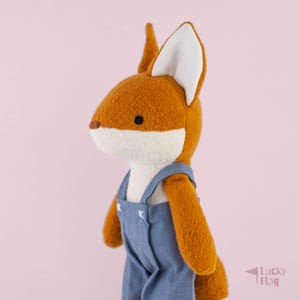 May include: A plush fox toy with textured orange fur and a white muzzle, ears, and chest. The fox is wearing blue denim-style overalls with white stitched details at the straps. The toy has small black eyes and a brown nose, presented in profile.