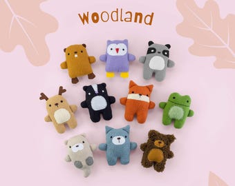 May include: A collection of ten small plush animal toys arranged in a circle on a light pink background. The text woodland is printed at the top and Lucky Flag at the bottom. The plushies include a beaver, owl, raccoon, deer, skunk, fox, frog, otter, cat, and bear.