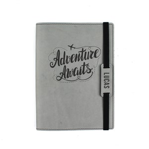 Puede incluir: Cuaderno de cuero gris con cierre elástico negro y una etiqueta de nombre personalizada que dice "LUCAS". El cuaderno tiene un diseño grabado que dice "Adventure Awaits" con un pequeño gráfico de avión.