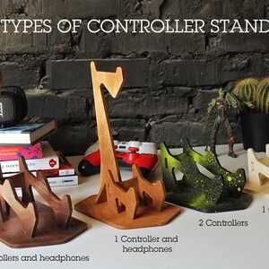 May include: Three different types of wooden controller stands. The first stand is brown and holds two controllers and headphones. The second stand is natural wood and holds one controller and headphones. The third stand is painted green and holds two controllers.
