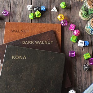 K&ouml;nnte beinhalten: Drei quadratische Holzbretter gestapelt mit den eingravierten Beschriftungen WALNUT, DARK WALNUT und KONA auf den Oberfl&auml;chen. Die Bretter sind von verschiedenen bunten polyedrischen W&uuml;rfeln und drei kleinen Glasgef&auml;&szlig;en mit Korkdeckeln umgeben.