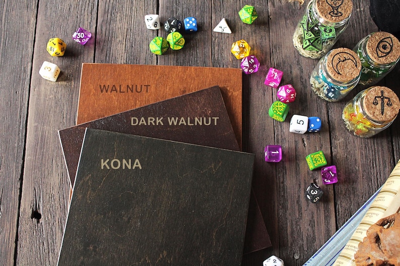 K&ouml;nnte beinhalten: Drei quadratische Holzbretter gestapelt mit den eingravierten Beschriftungen WALNUT, DARK WALNUT und KONA auf den Oberfl&auml;chen. Die Bretter sind von verschiedenen bunten polyedrischen W&uuml;rfeln und drei kleinen Glasgef&auml;&szlig;en mit Korkdeckeln umgeben.