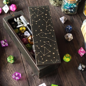 May include: A wooden box with a black and gold starry sky design. The box is open and contains a variety of dice, a pen, and other small items. The box is sitting on a wooden table.