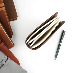 May include: A stack of brown leather bound journals with elastic closures, a single open journal with blank pages, and a green pen on a white background.