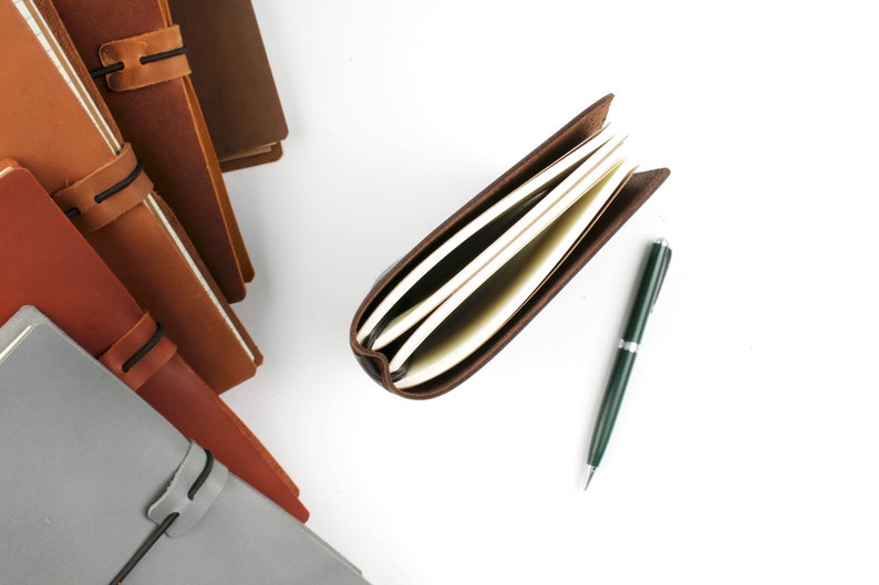 May include: A stack of brown leather bound journals with elastic closures, a single open journal with blank pages, and a green pen on a white background.