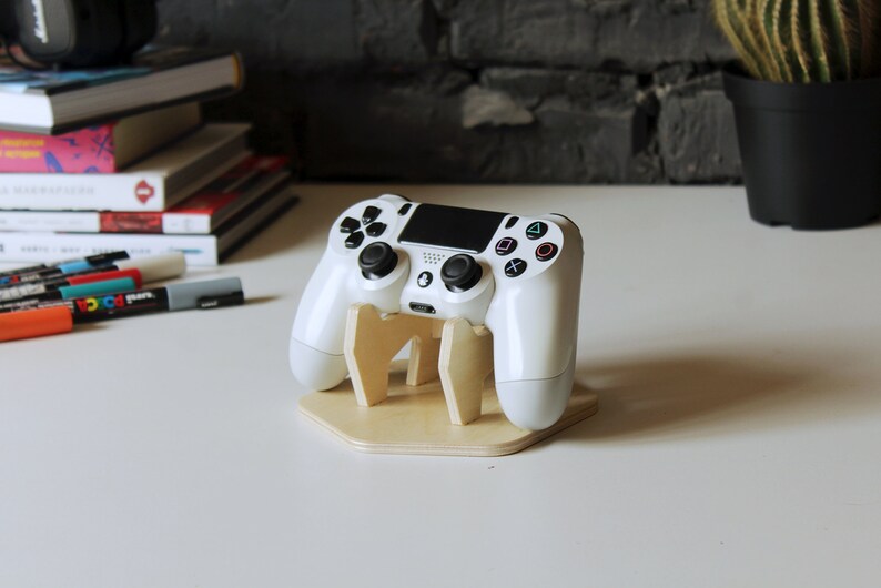 May include: A white PlayStation 4 controller is displayed on a wooden stand. The stand is a light brown color and has a hexagonal base.
