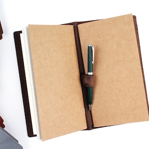 May include: A brown leather bound journal with a pen holder and a green pen inserted. The journal is open to blank pages.