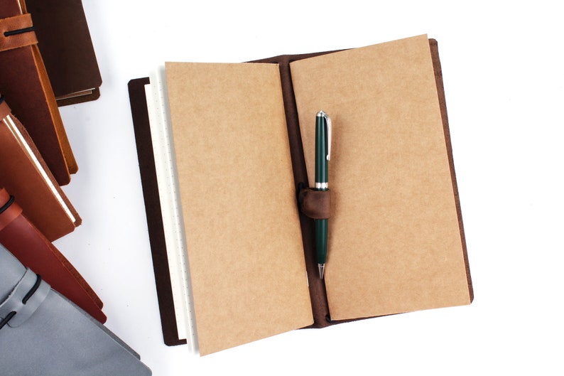 May include: A brown leather bound journal with a pen holder and a green pen inserted. The journal is open to blank pages.