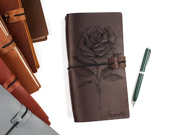 May include: A brown leather journal with a rose design and the name "Janelle" engraved on the cover. A green pen is lying next to the journal.