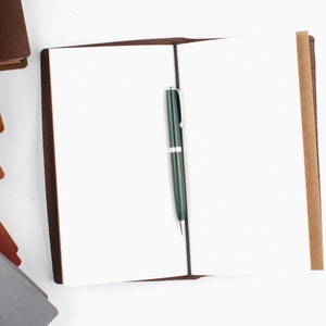 May include: A brown leather bound journal with blank pages and a green pen lying open on a white surface.
