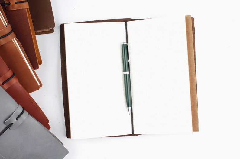 May include: A brown leather bound journal with blank pages and a green pen lying open on a white surface.