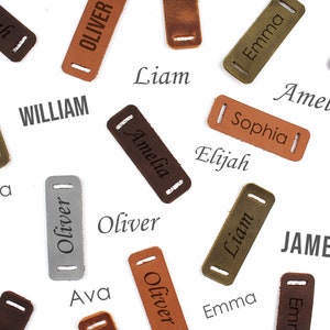 Puede incluir: Una colecci&oacute;n de etiquetas de nombre de cuero con varios nombres, incluyendo Elijah, Liam, James, Oliver, William, Amelia, Sophia, Ava y Emma. Las etiquetas son marrones, verdes, grises y plateadas.