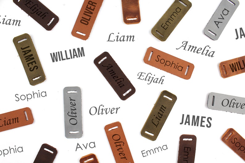 Puede incluir: Una colecci&oacute;n de etiquetas de nombre de cuero con varios nombres, incluyendo Elijah, Liam, James, Oliver, William, Amelia, Sophia, Ava y Emma. Las etiquetas son marrones, verdes, grises y plateadas.