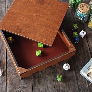 K&ouml;nnte beinhalten: Eine quadratische W&uuml;rfelbox aus Holz mit abnehmbarem Deckel, dunkelbraun gebeizter Au&szlig;enseite und einem mit tiefrotem Filz ausgekleideten Innenraum. Die Box ist umgeben von verschiedenen mehrseitigen Spielw&uuml;rfeln, Gl&auml;sern und einem Totenkopf.