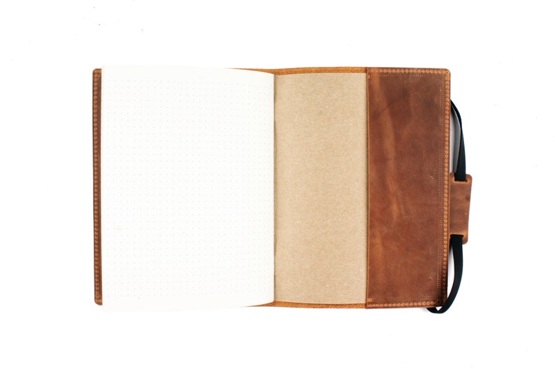 Puede incluir: Una cubierta de cuaderno de cuero marr&oacute;n con una correa negra. La cubierta est&aacute; abierta para revelar un cuaderno blanco punteado en blanco.