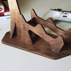 May include: A dark brown wooden stand with two slots for holding video game controllers. The stand has a unique, geometric design and a wide base for stability.