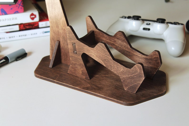 May include: A dark brown wooden stand with two slots for holding video game controllers. The stand has a unique, geometric design and a wide base for stability.