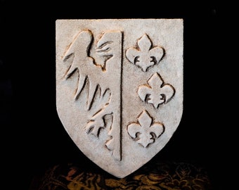 Op de afbeelding: Een wit keramisch schild met een gesneden ontwerp. De linkerzijde van het schild toont een gestileerde vogel met gespreide vleugels. De rechterzijde van het schild toont drie gestileerde fleurs-de-lis.