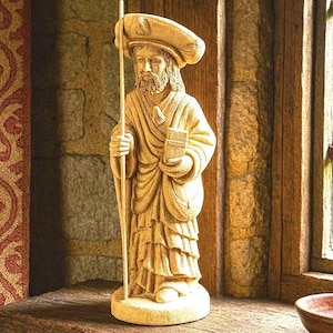 May include: A carved wooden statue of a robed figure with a hat, holding a staff and a book. The statue is light brown and stands on a wooden shelf near a window. The figure has a beard and is wearing a hat.