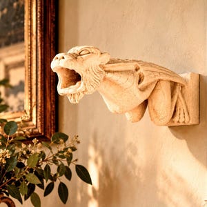 Puede incluir: Una escultura de pared de piedra beige que representa una gárgola agachada con la boca abierta, las alas plegadas contra la espalda y detalles texturizados. La criatura está montada horizontalmente sobre una superficie plana y sobresale.