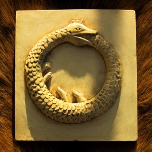 May include: A white ceramic tile featuring a raised relief sculpture of a snake biting its own tail, forming a circle. The snake is detailed with scales and has a crown on its head.