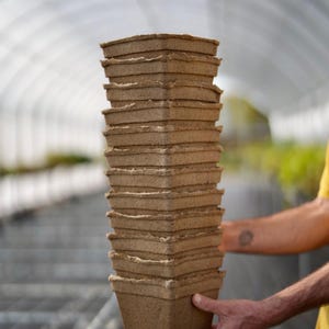 May include: A stack of brown biodegradable pots for planting, made from recycled paper pulp. The pots are stacked in a person's hand.