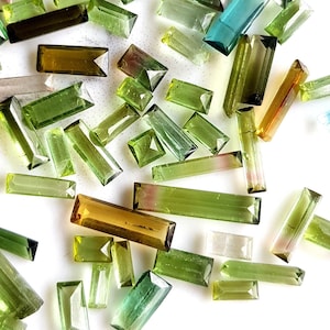 May include: A collection of green, blue, and brown tourmaline gemstones cut into rectangular shapes. The gemstones are scattered on a white surface.
