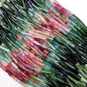 May include: A strand of multi-colored tourmaline beads. The beads are long and thin, and they are arranged in a gradient from green to pink. The beads are perfect for making jewelry.
