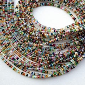 Può includere: Fili di perline di pietre preziose multicolori. Le perline sono un mix di colori, tra cui blu, verde, giallo, rosso e trasparente. Le perline sono infilate con un filo bianco.