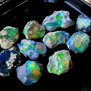 May include: A collection of rough opal stones with a variety of iridescent colours, including green, blue, and orange. The stones are set against a black background.