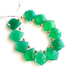 Puede incluir: Una hebra de cuentas de piedras preciosas verdes con detalles dorados. Las cuentas son facetadas y tienen forma de lágrima. La hebra está enhebrada en un alambre de plata.