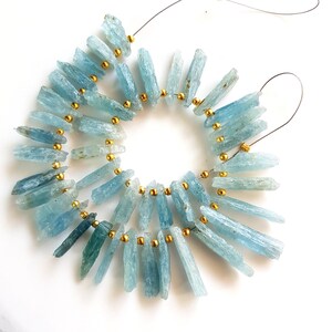 May include: A strand of light blue aquamarine crystal beads with gold spacers. The beads are long and narrow, resembling shards of crystal. The strand is strung on a wire and is ready to be used for jewellery making.