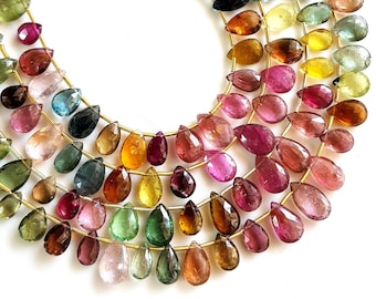 May include: Strands of colorful, teardrop-shaped gemstones in shades of pink, green, yellow, and brown. The gemstones are strung on gold-colored wire, creating a layered effect. The gemstones are arranged on a white surface.