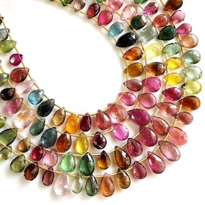 May include: Strands of colorful, teardrop-shaped gemstones in shades of pink, green, yellow, and brown. The gemstones are strung on gold-colored wire, creating a layered effect. The gemstones are arranged on a white surface.