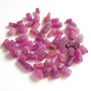 May include: A collection of small, rough, pink ruby gemstones. The rubies are various shapes and sizes, and have a natural, unpolished look.