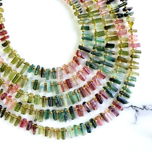 May include: A multi-strand necklace made of green, pink, and black tourmaline beads. The beads are faceted and have a gold-toned metal spacer between each bead.