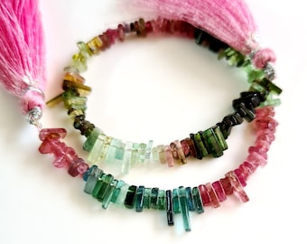 May include: A strand of multi-colored tourmaline beads with pink tassels at each end. The beads are faceted and have a variety of shapes and sizes. The colors of the beads include pink, green, and black.