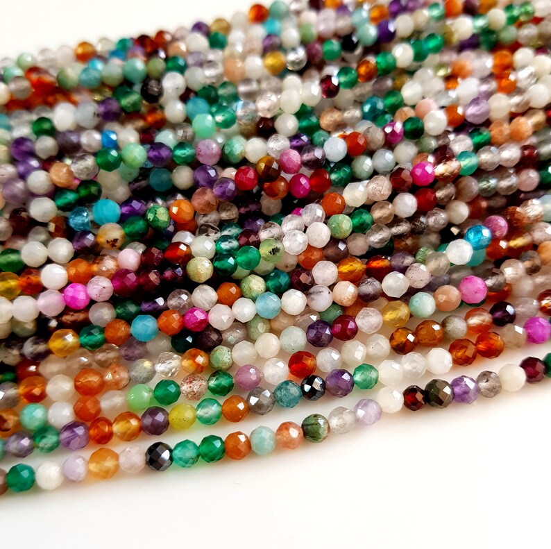 May include: A close-up view of a collection of faceted gemstone beads in a variety of colors. The beads are strung together, showcasing a spectrum of hues including green, purple, orange, and pink. The beads are small and round, with a sparkling, polished finish.