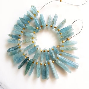 May include: A strand of aquamarine gemstone beads strung on wire with gold spacers. The beads are long and slender, with a light blue colour.