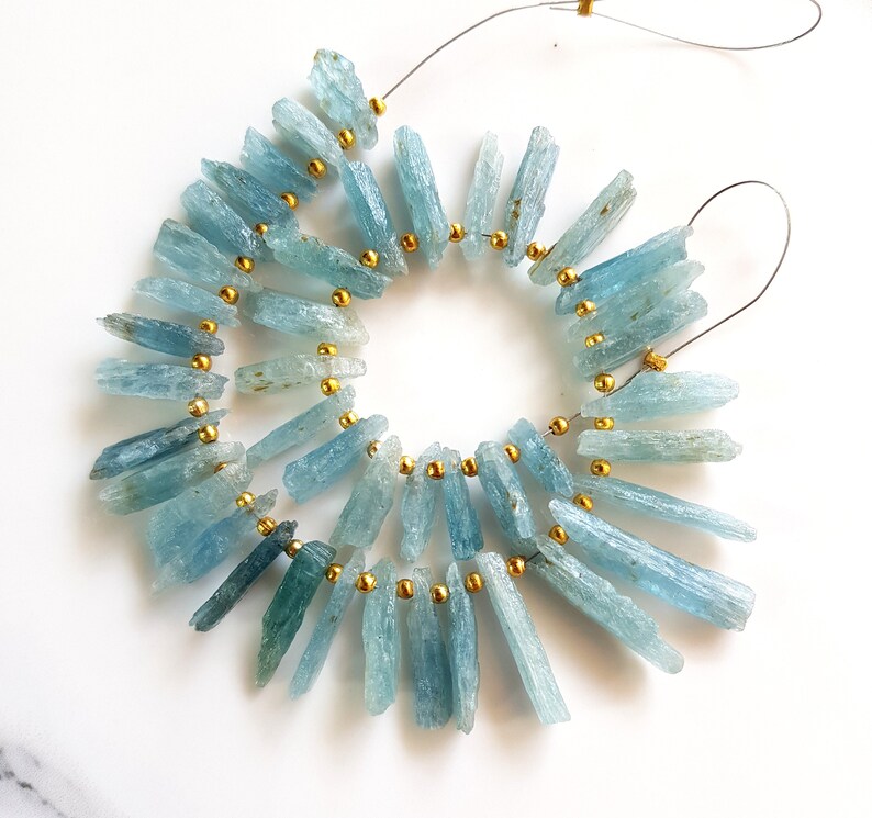 May include: A strand of aquamarine gemstone beads strung on wire with gold spacers. The beads are long and slender, with a light blue colour.
