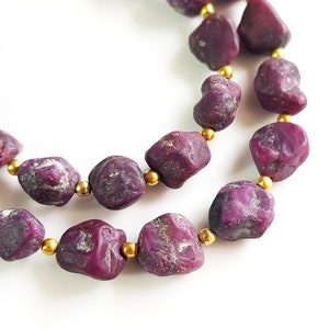 Könnte beinhalten: Eine Kette aus tiefvioletten Edelsteinperlen mit goldenen Zwischenstücken. Die Perlen sind unregelmäßig geformt und haben eine raue, natürliche Textur.